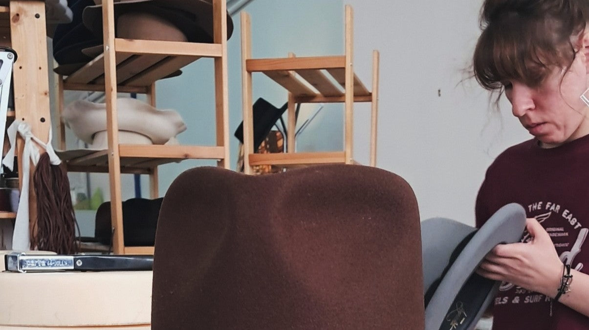 Brown custom felt hat in the foreground of a workshop, with unfinished hats on wooden shelves and a person inspecting a grey hat in the background.