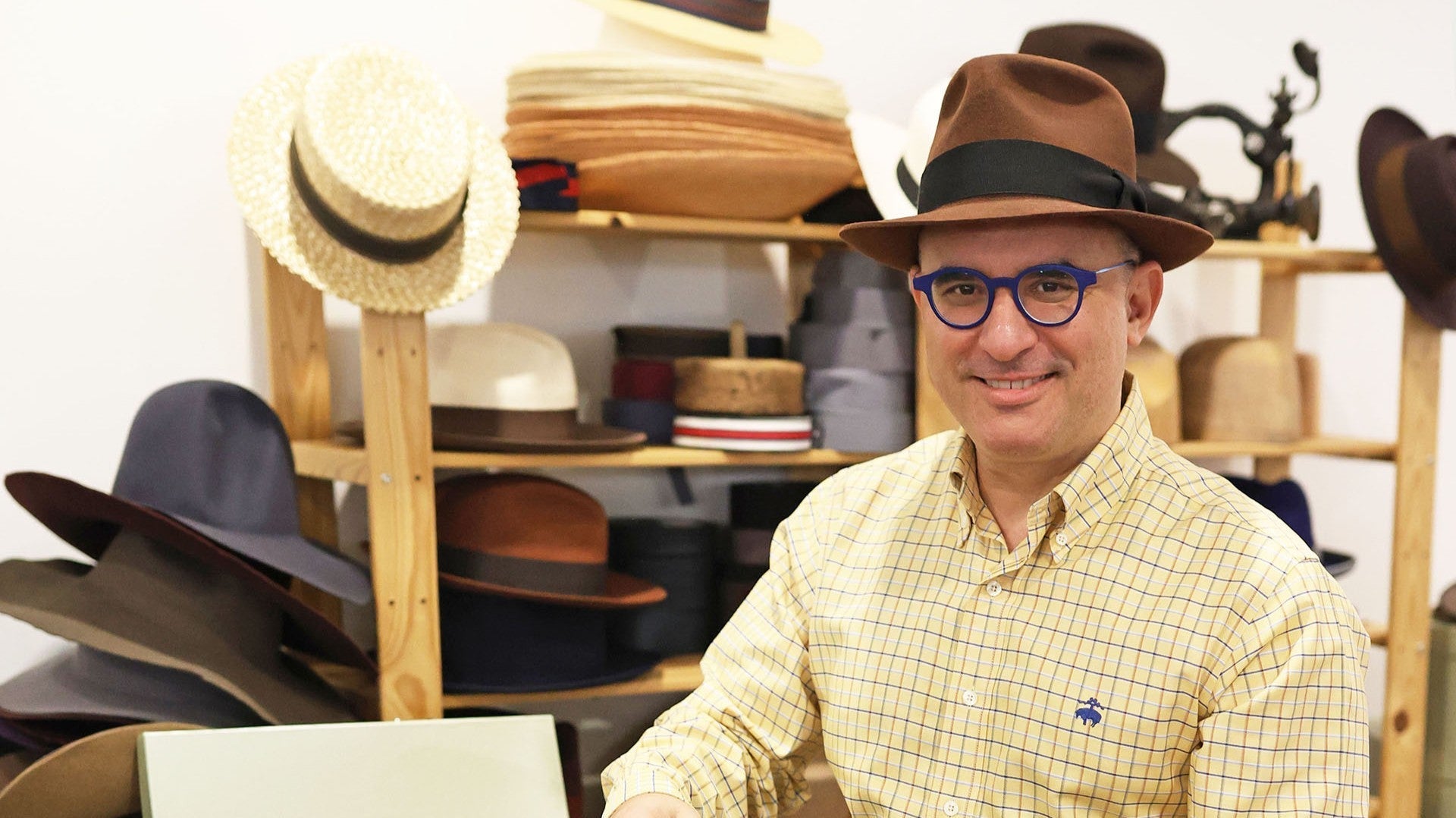 Hat artisan in a workshop brushing a black top hat, surrounded by hat-making tools, wooden blocks, and various handcrafted hats displayed on shelves.