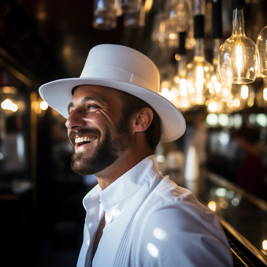 A joyful man wearing a pristine white Agnoulita hat, lit by ambient golden lighting, which accentuates his radiant smile. He stands near a bar adorned with illuminated light bulbs, reflecting the elegance and warmth of the Sunset Silhouettes theme.