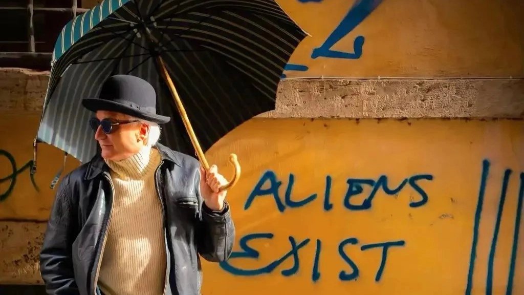 playful photo of a man with casual and a classic bowler hat on. He is holding a colorful umbrella. the graffiti on the wall reads "Aliens Exist"