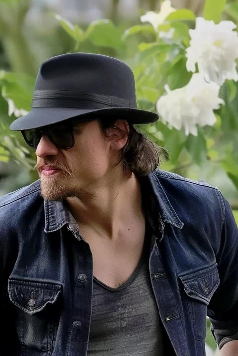 Man in a casual denim jacket wearing a stylish small brim black fedora, seated in a lush garden setting.