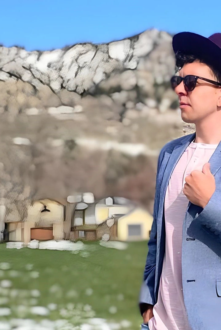 A fashion-forward young man in sunglasses and a stylish fedora with a feather accent stands confidently in a field, with a backdrop of snowy mountains and quaint houses.