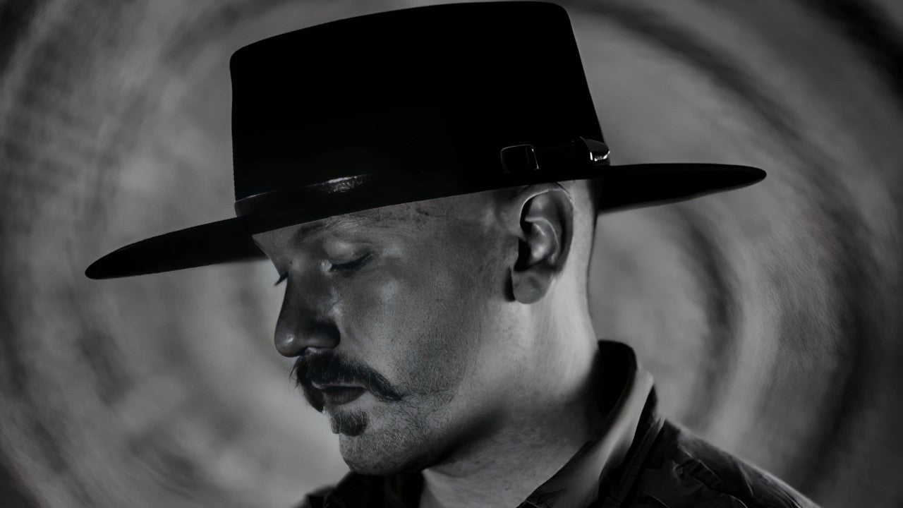 A man in a dramatic black and white setting wears a flat brim cowboy hat, capturing a moment of deep contemplation and rugged style.