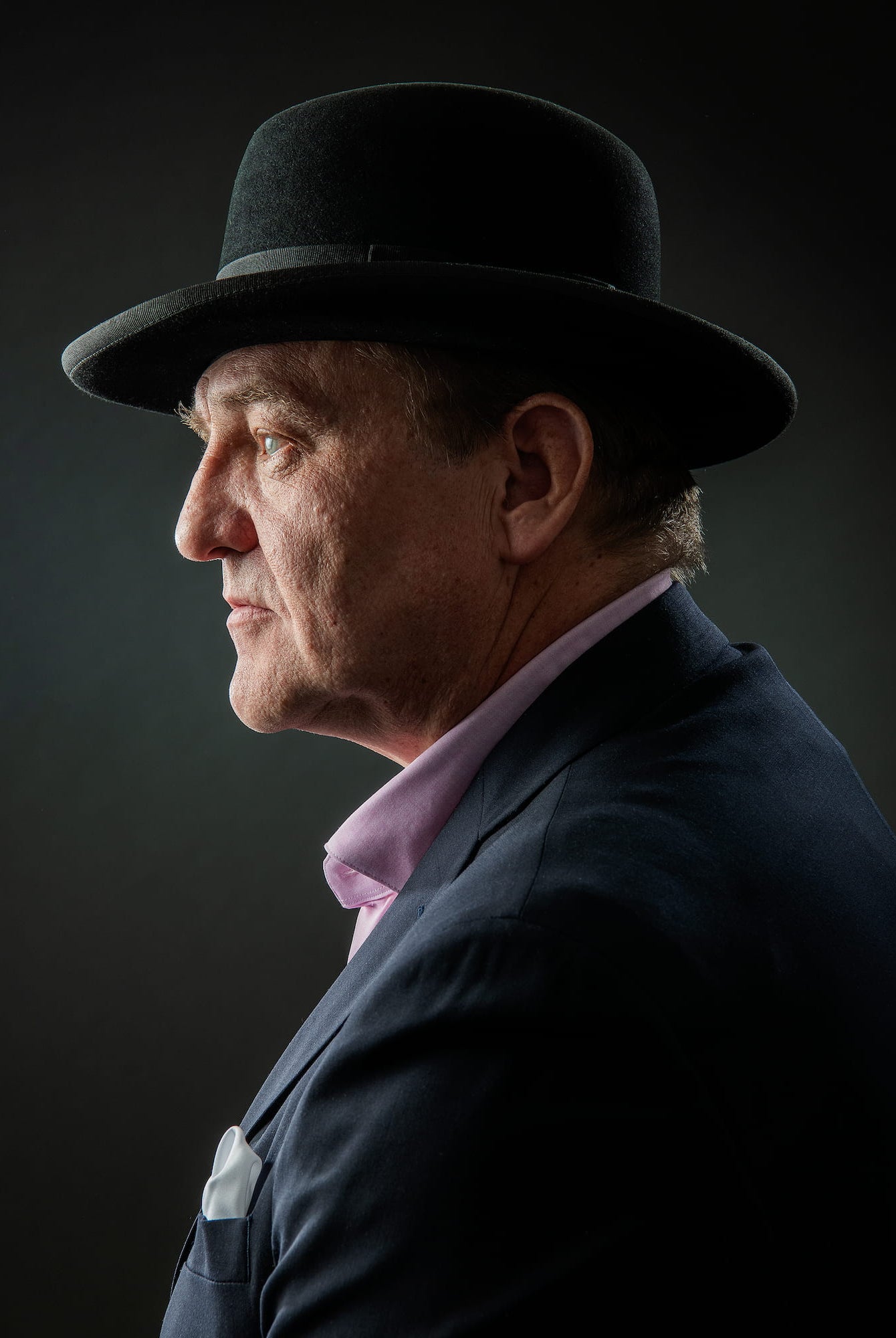 Man wearing a dark suit, light shirt, and black hat against a dark background