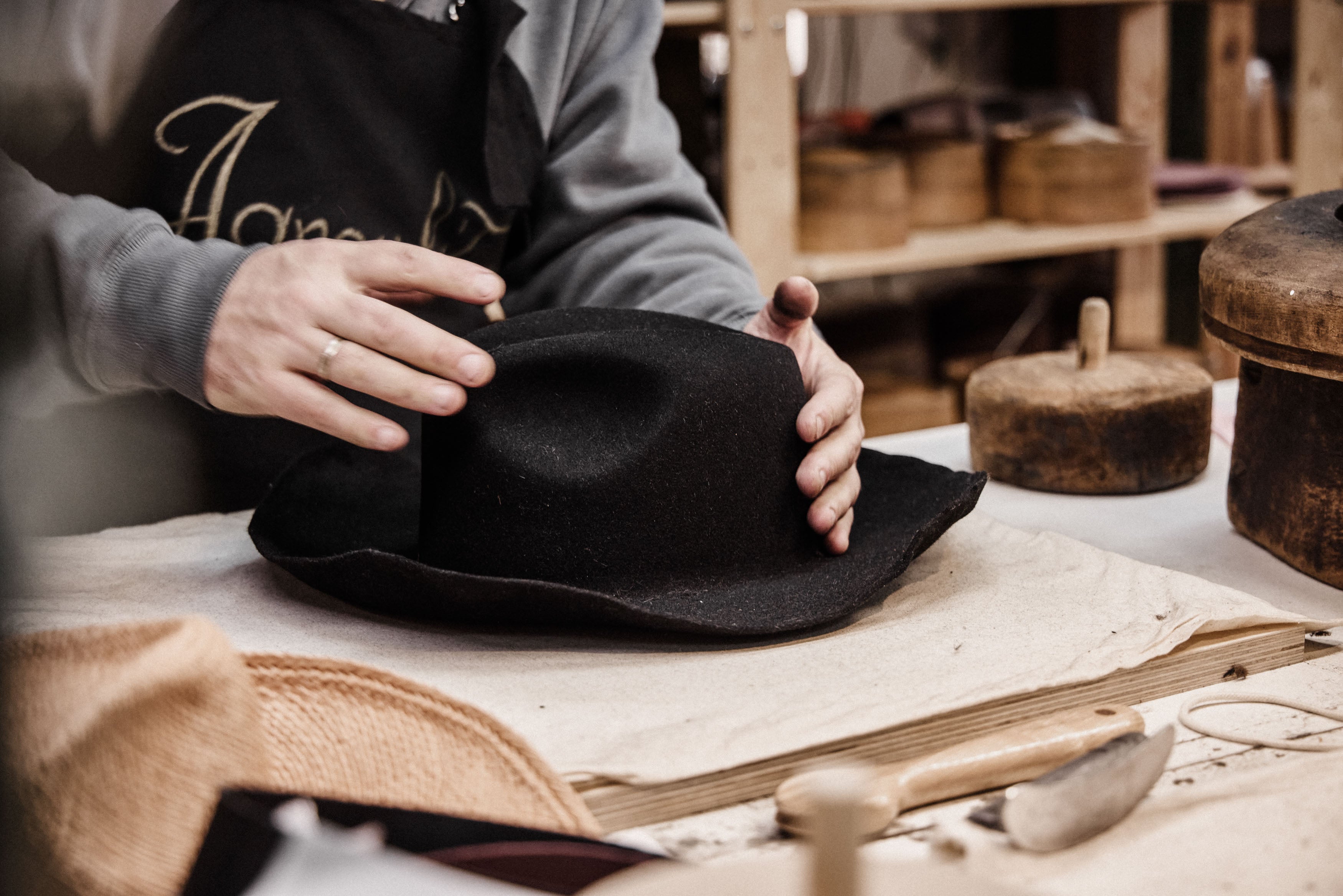 Person wearing a black apron with gold text, holding a black hat in a workshop setting.