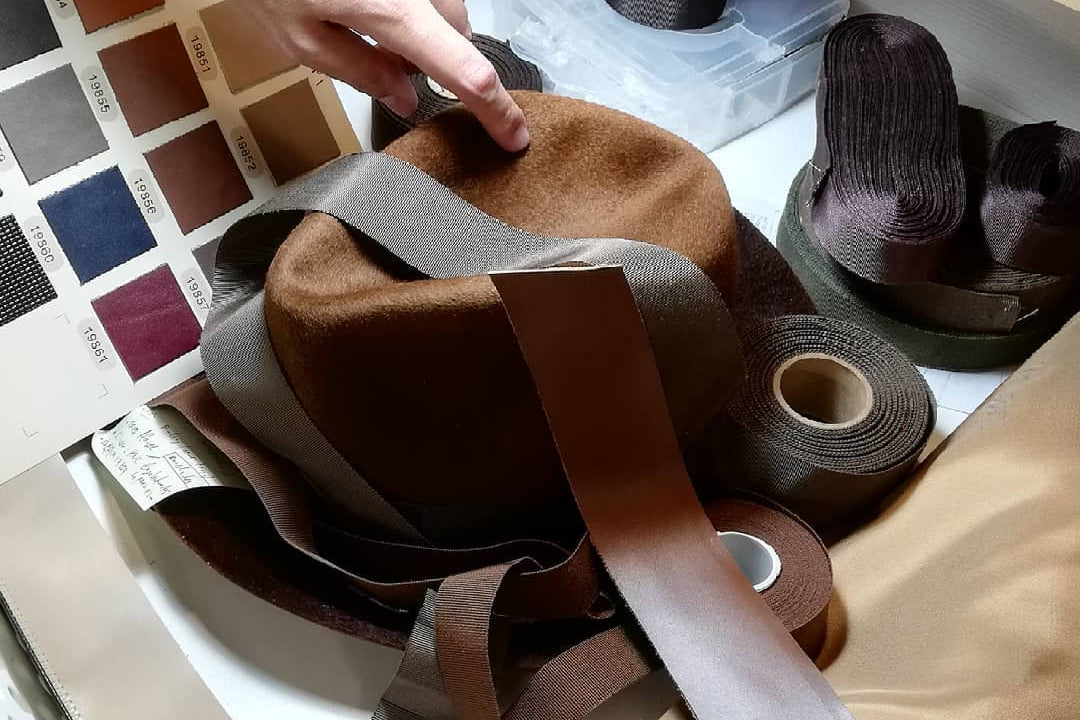 Person selecting a color swatch for a brown hat and ribbon on a table with fabric and thread.