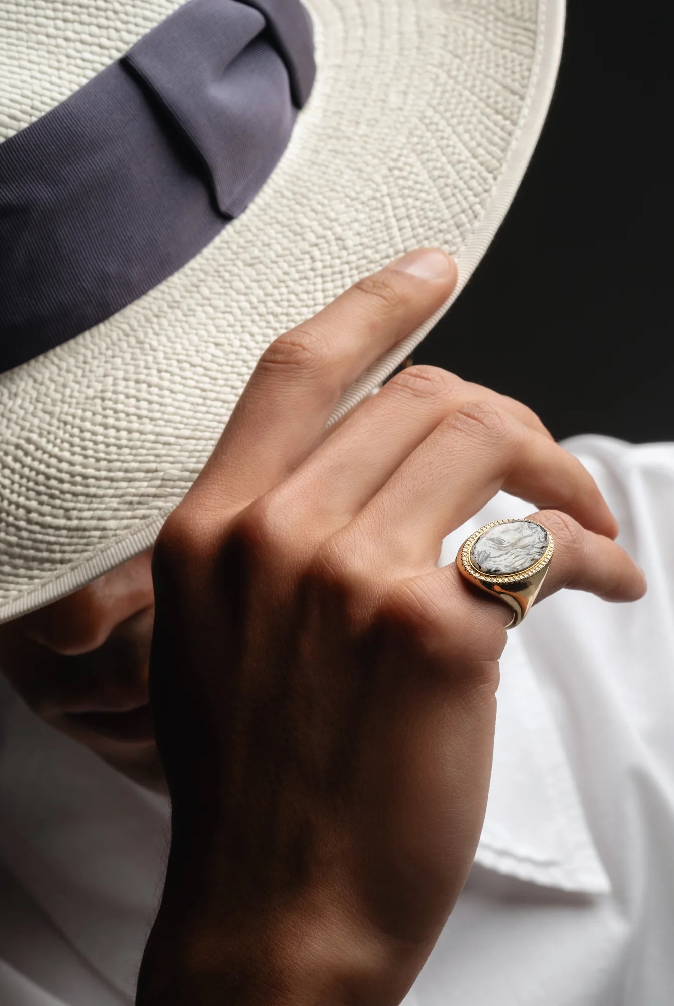 Hand wearing a gold ring with a diamond, holding a white hat against a dark background