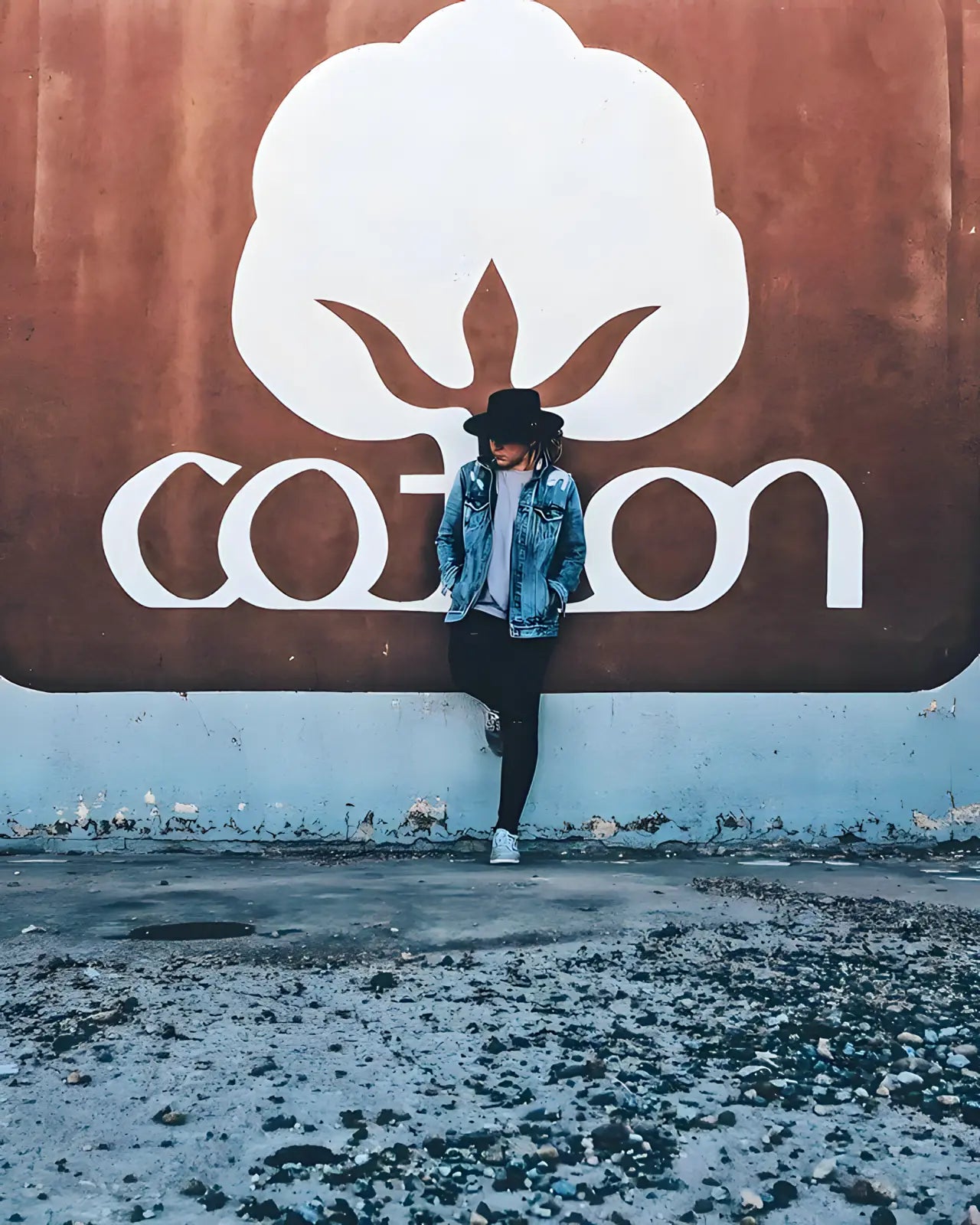A young person wearing a wide-brimmed black hat and a denim jacket stands in front of a large cotton logo, symbolizing commitment to sustainable fashion.