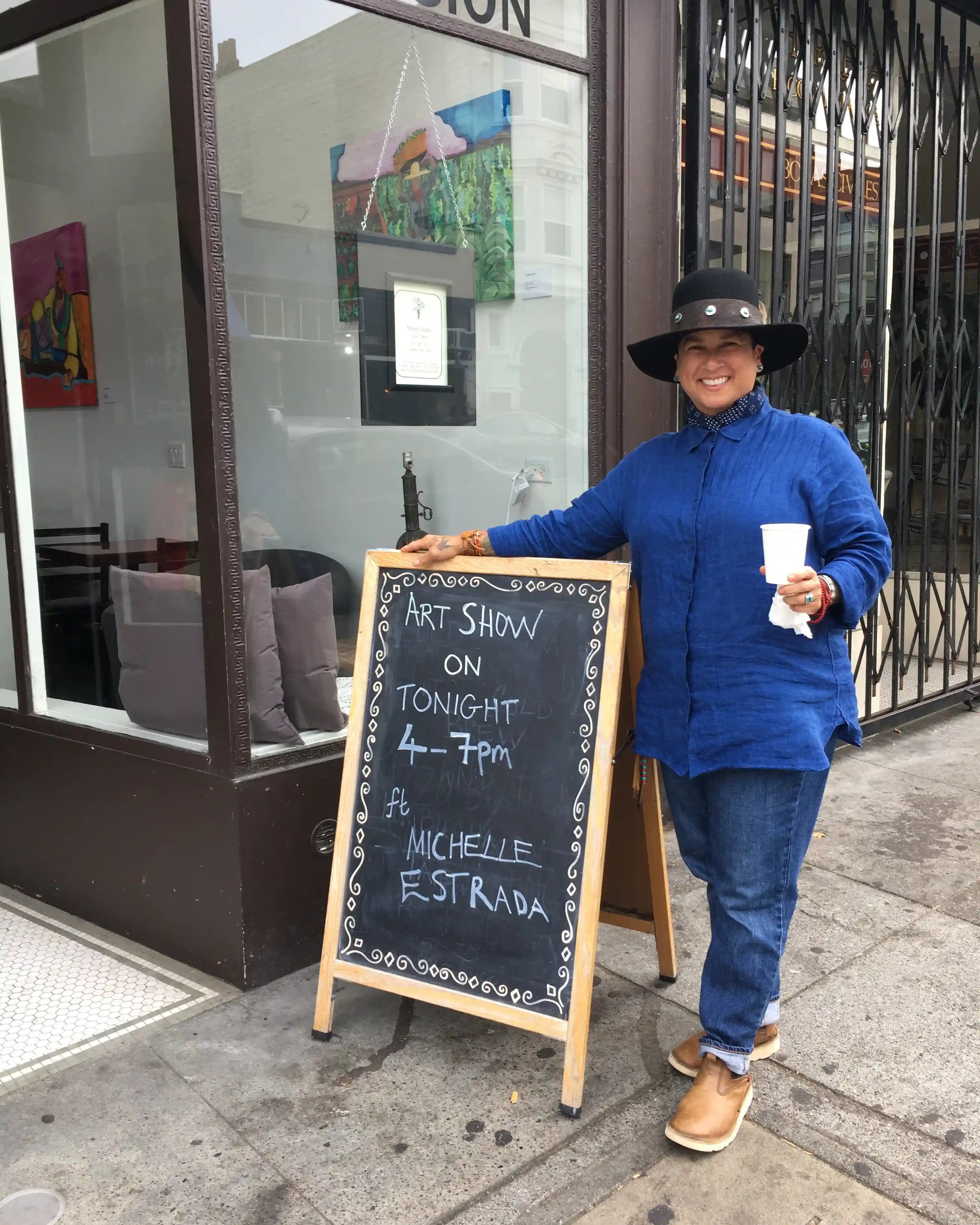 A cheerful woman in a denim outfit and a sustainable handmade black hat stands outside an art gallery, promoting an art show next to a chalkboard sign