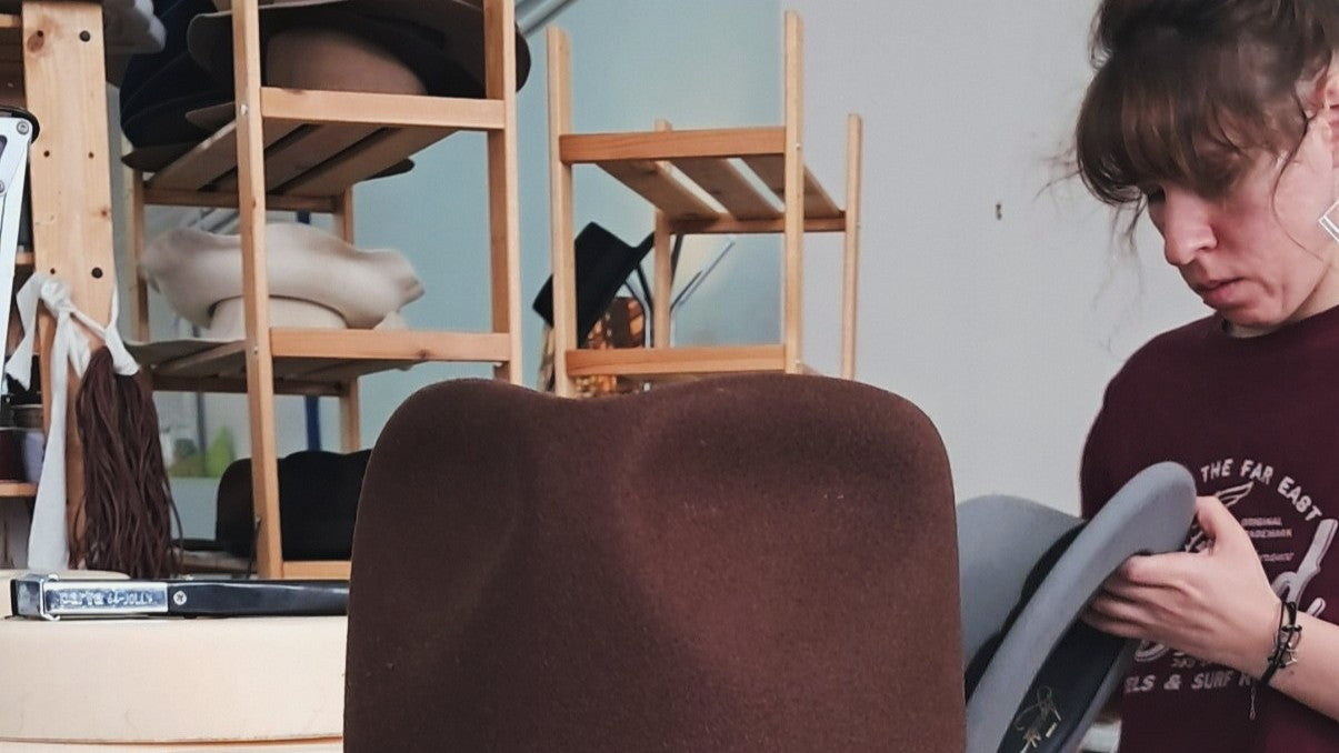 Brown custom felt hat in the foreground of a workshop, with unfinished hats on wooden shelves and a person inspecting a grey hat in the background.