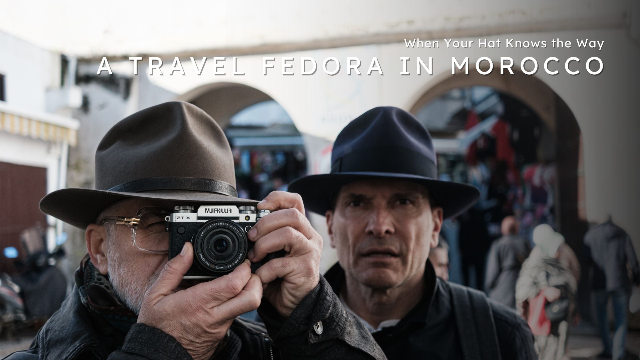Blog banner showing two people in fedoras in a busy Moroccan street, one lifting a camera, with the title text ‘A Travel Fedora in Morocco – When Your Hat Knows the Way’ overlaid at the top.
