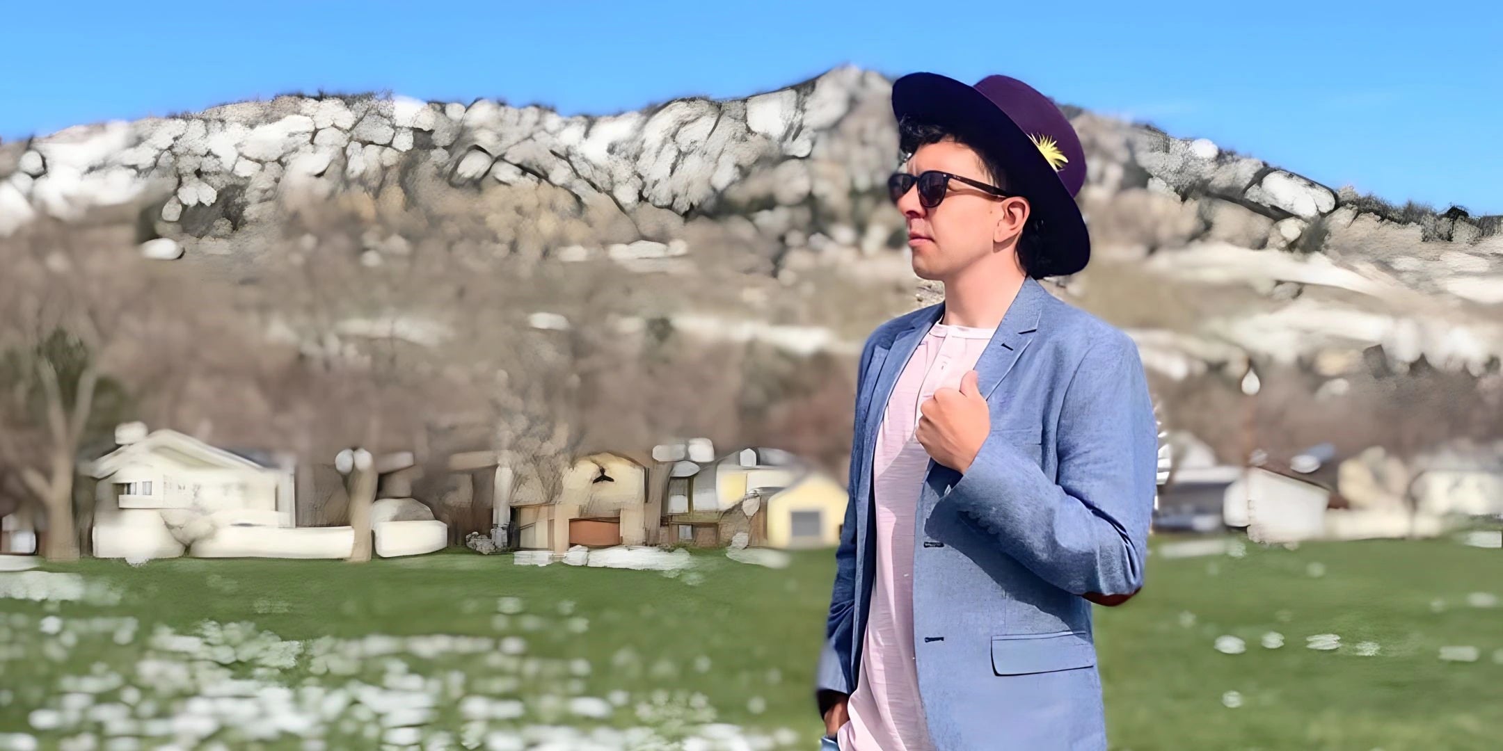 A fashion-forward young man in sunglasses and a stylish fedora with a feather accent stands confidently in a field, with a backdrop of snowy mountains and quaint houses.