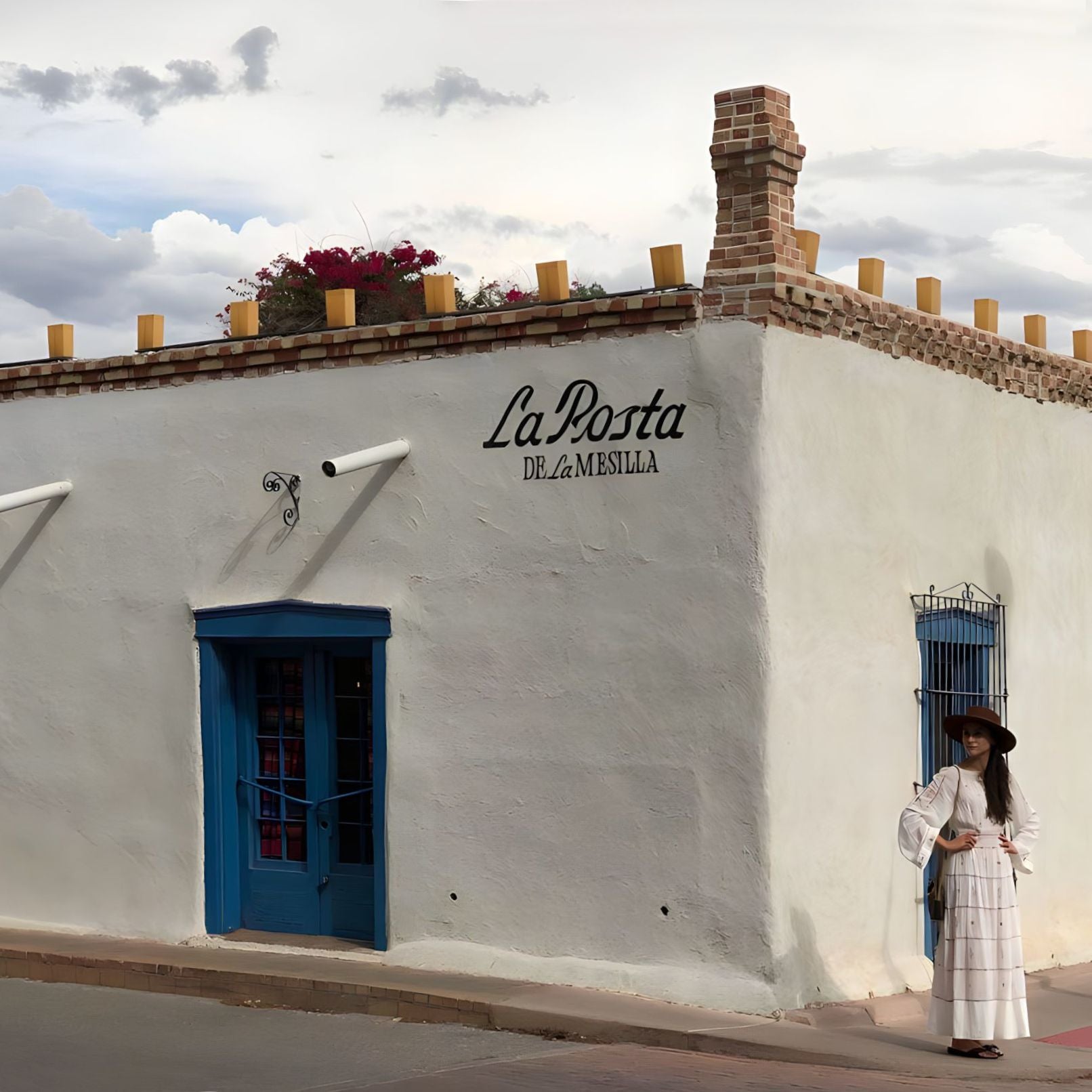 A woman in a stylish women's western hat stands in front of La Posta de La Mesilla, her elegance accentuating the rustic charm of the historic setting.