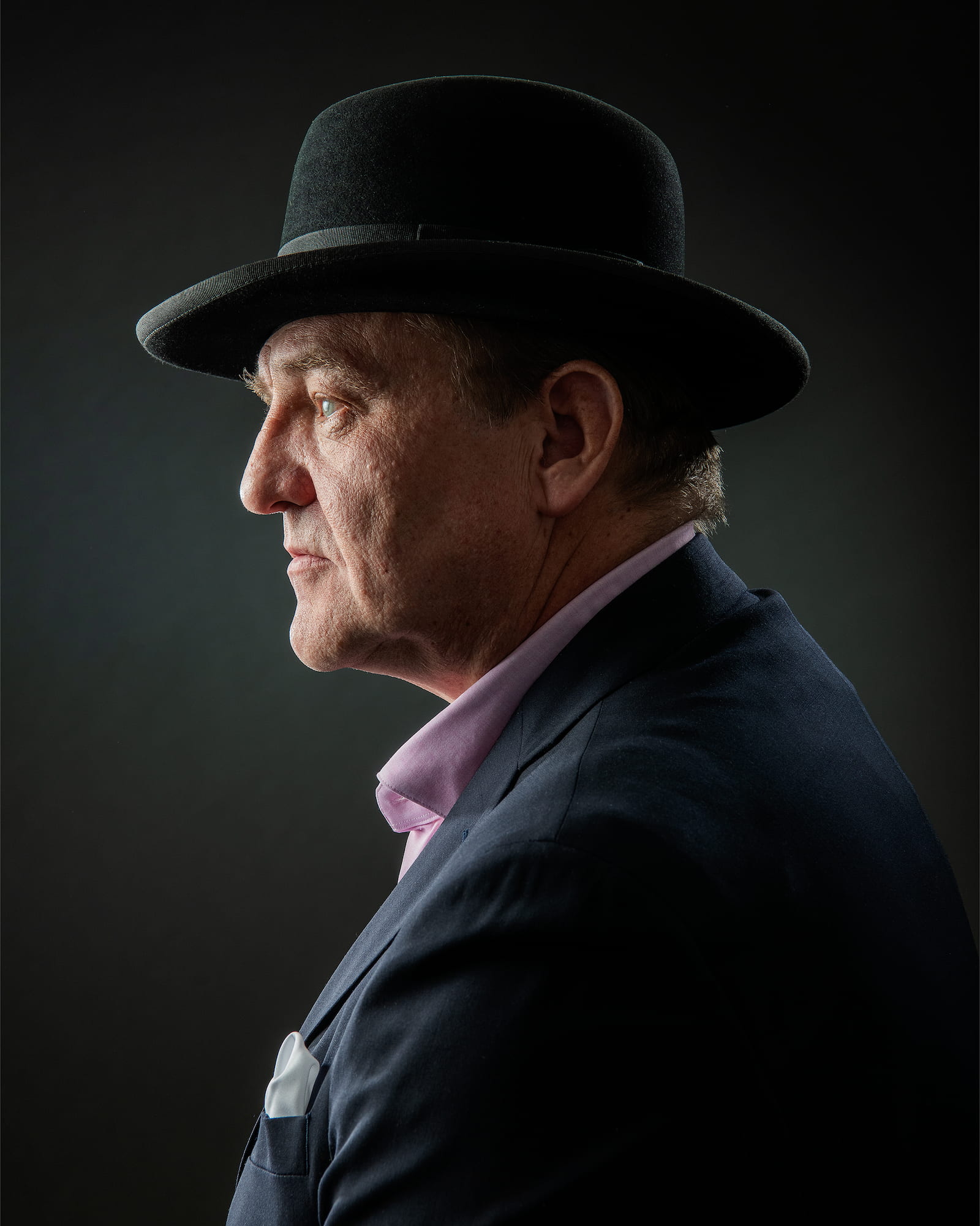 Man wearing a dark suit, light shirt, and black hat against a dark background