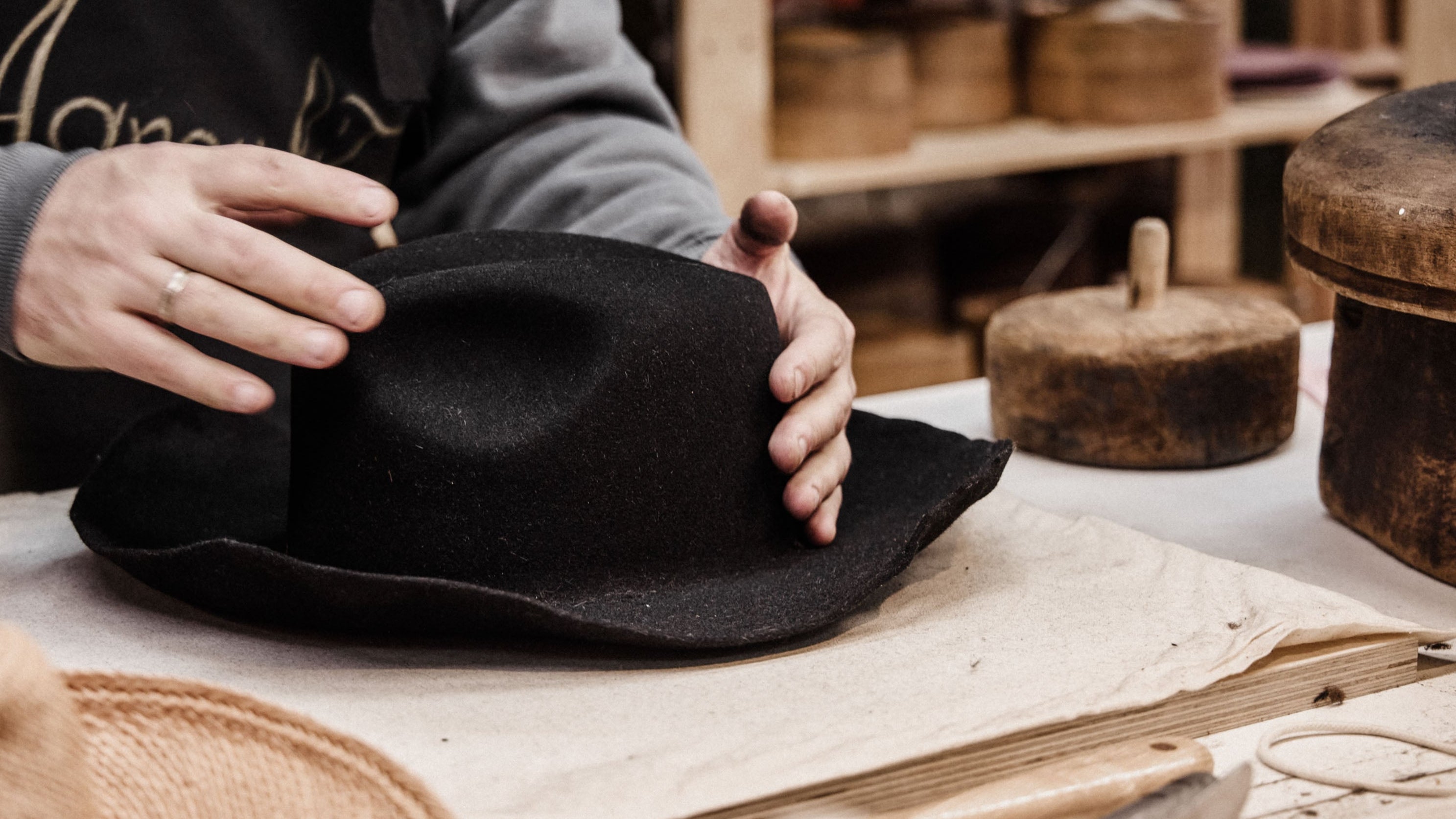 Person wearing a black apron with gold text, holding a black hat in a workshop setting.