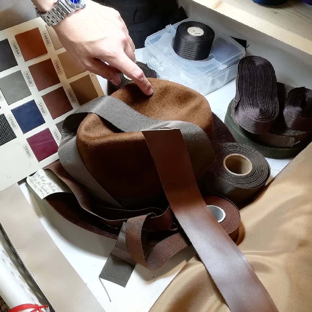 Person selecting a color swatch for a brown hat and ribbon on a table with fabric and thread.