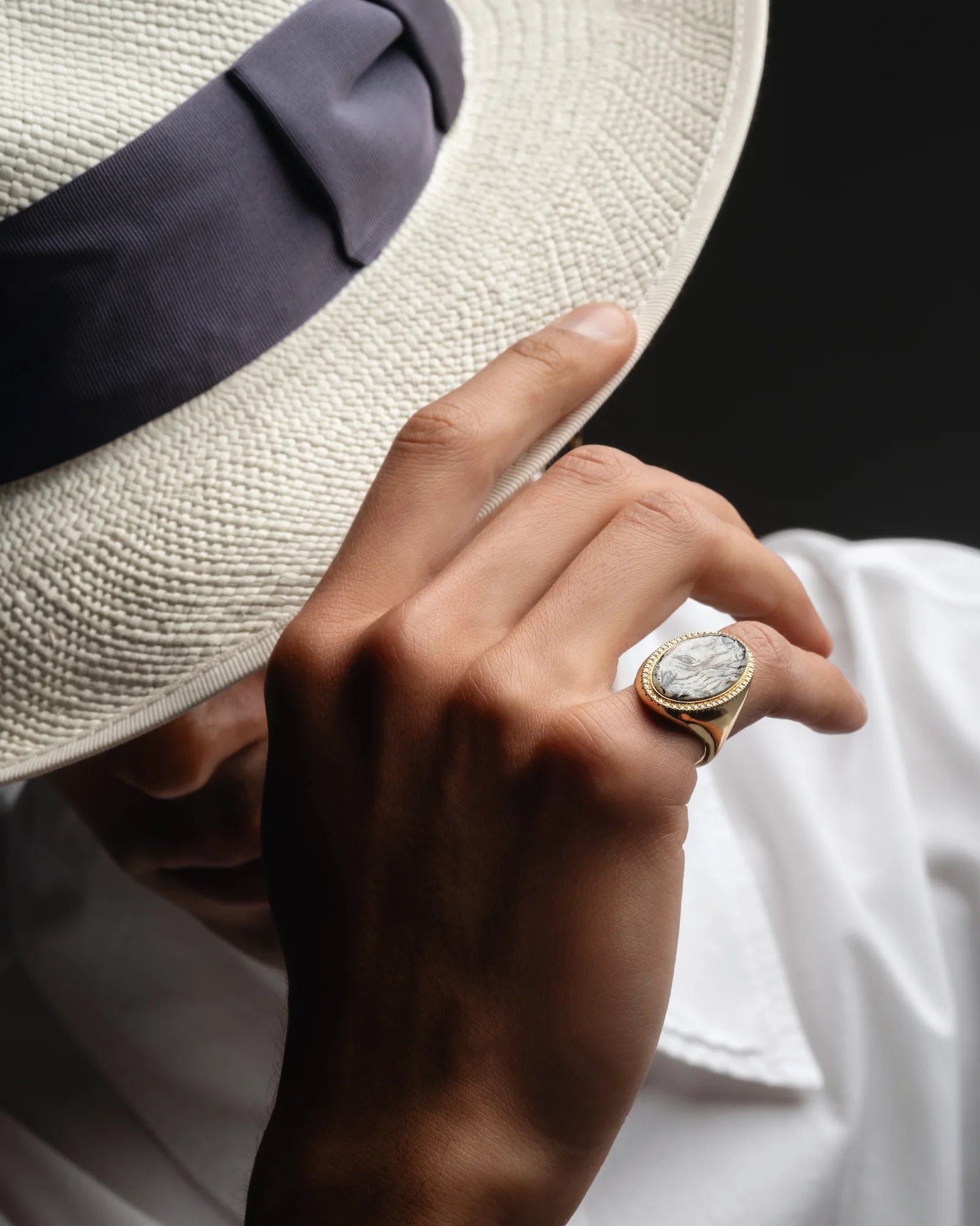 Hand wearing a gold ring with a diamond, holding a white hat against a dark background