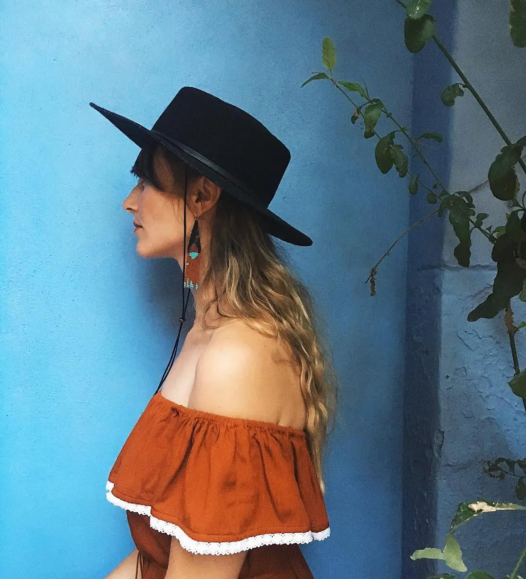 Side view of a woman in a western boho outfit posing against a blue wall, wearing her Agnoulita Gambler hat