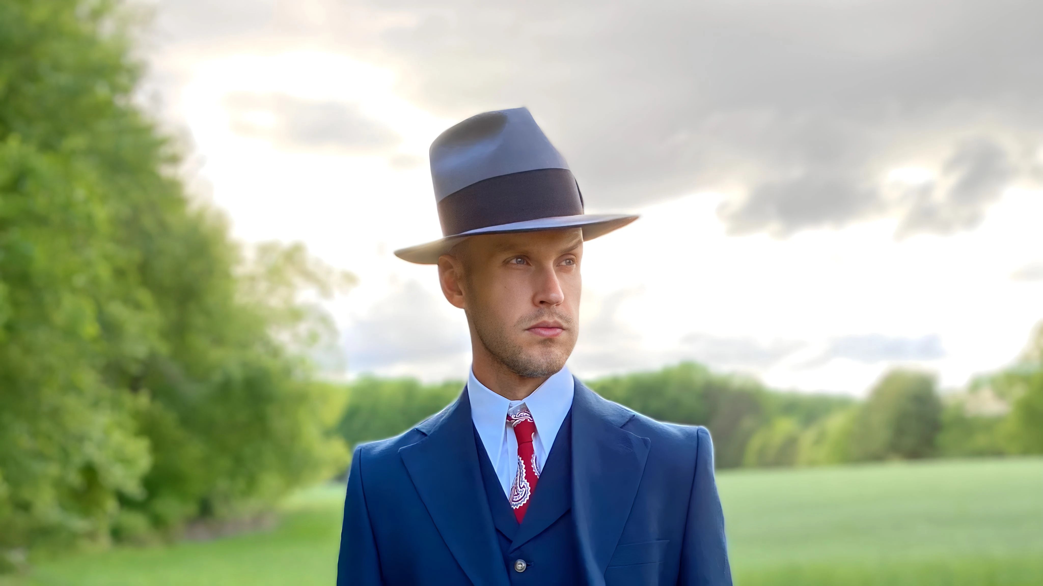 Well-dressed gentleman standing under a cloudy pastel sky, wearing his new custom beaver fedora from Agnoulita Hats.