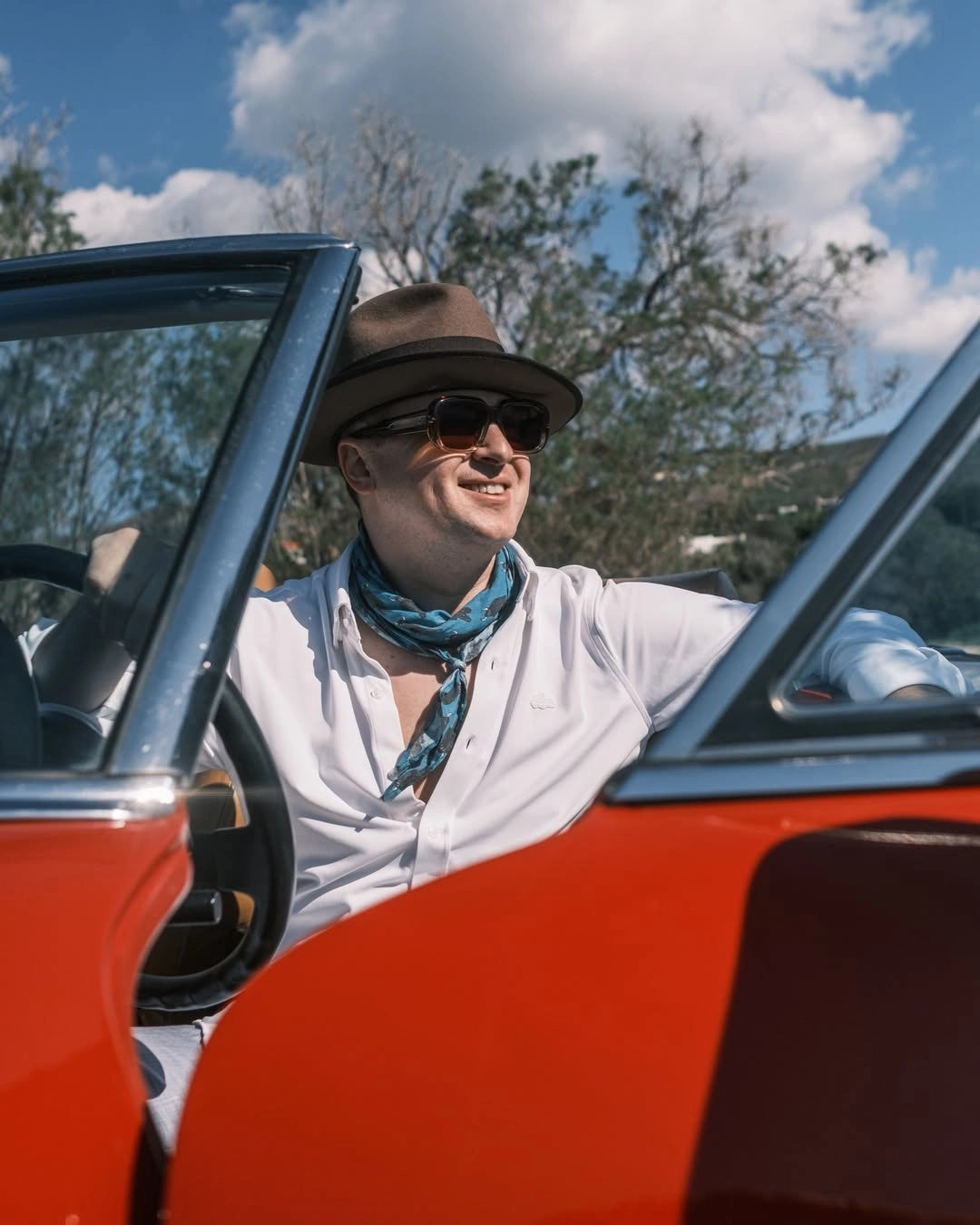 Man wearing brown Agnoulita fedora and sunglasses leaning out of vintage orange convertible car against blue sky