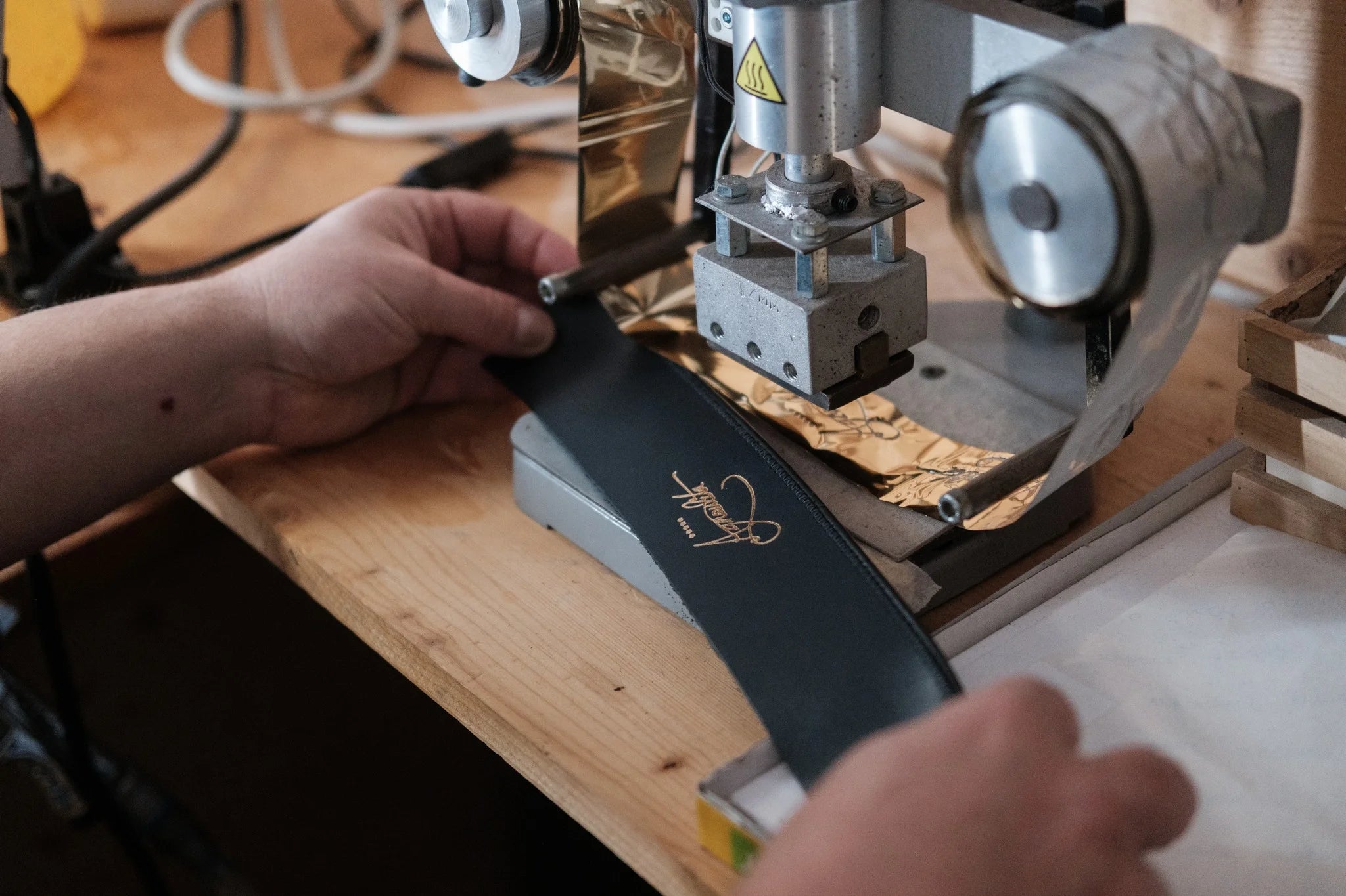 Close-up of a craftsman's hands holding a black leather hat sweatband under a hot stamping machine, pressing a gold foil "Agnoulita Hats" logo onto the material with a roll of gold foil visible on a wooden workshop workbench.